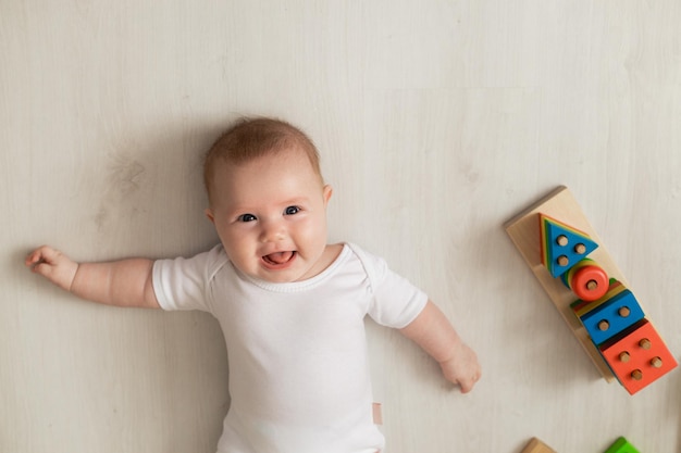 Cheerful newborn baby in a white bodysuit lies on his back on floor and plays with educational toys