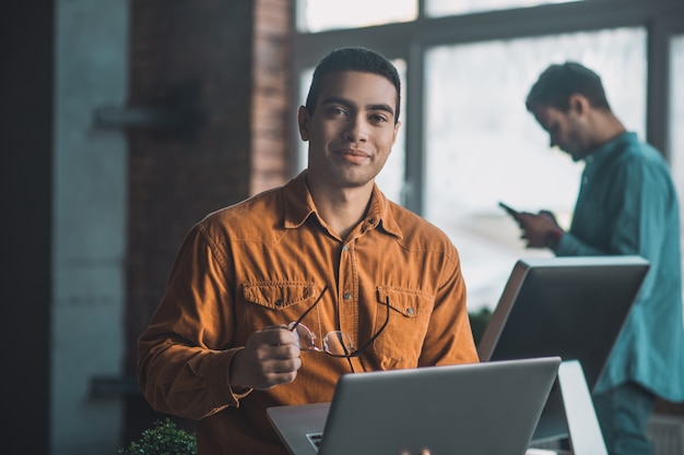 Cheerful nice man holding his eyeglasses in the hand while being distracted from his work