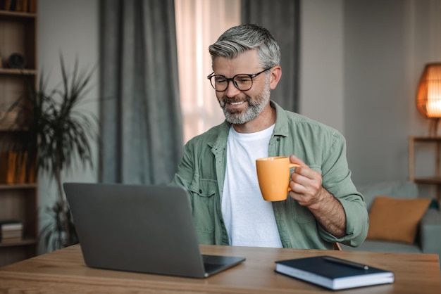 Cheerful old caucasian man with beard in glasses typing on laptop at workplace drinking favorite drink in room