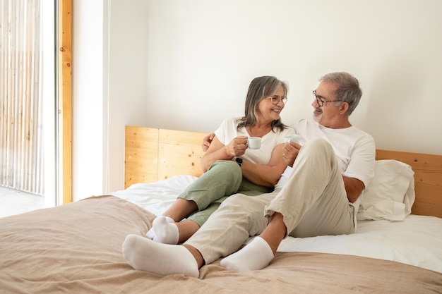 Cheerful old european man and lady have breakfast with cups of coffee enjoy free time sit on bed in