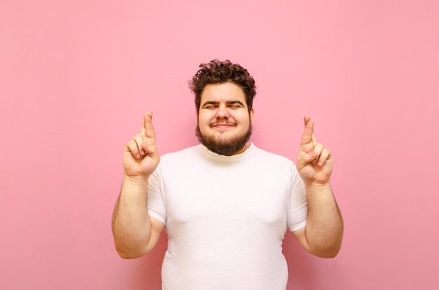 Cheerful overweight boy crossed fingers with closed eyes wearing white tshirt isolated