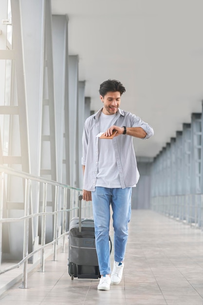 Cheerful passenger with luggage looking at watch at airport