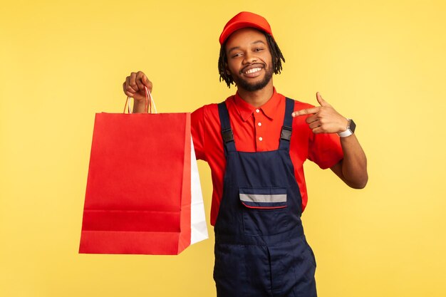 Cheerful positive delivery man in uniform pointing finger at shopping bags in his hand, fast delivery from store, service industry. Indoor studio shot isolated on yellow background.