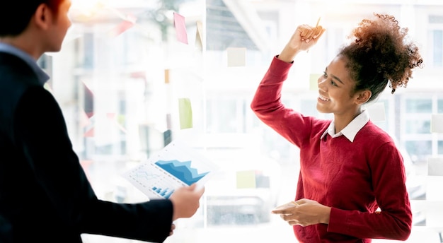 Cheerful positive ethnic freelancer looking at sticky note on the wall and holding arms up yelling celebrating victory achievement successfully completed project