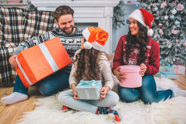 Cheerful and positive family sitting on floor and smiling. They look at each other. Father holds big box of present. Young woman has pink one while girl has blue box in hands. They look festive.