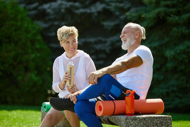 Cheerful positive smiling mature people man and woman sitting on bench outdoors with fitness mat