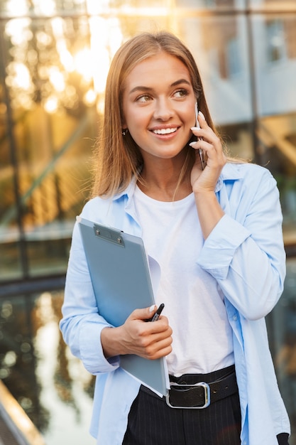 cheerful positive young business lady with clipboard posing outdoors near business center talking bymobile phone.