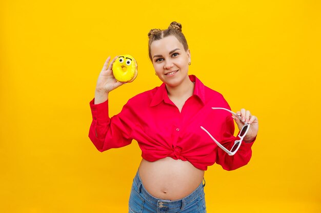 Cheerful pregnant woman holding a sweet yellow donut in her hand on a yellow background Expecting a child pregnancy and motherhood The concept of healthy and unhealthy food diet Junk food