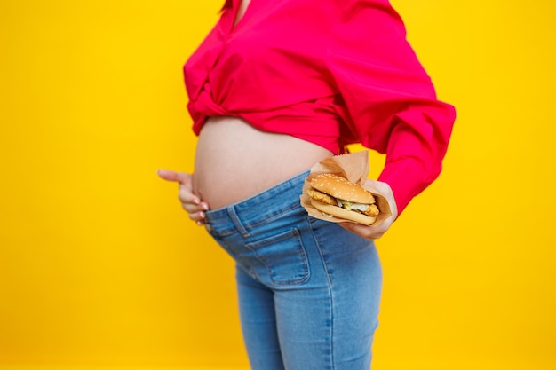 Cheerful pregnant woman in pink shirt holding hamburger over isolated yellow background with surprise and shocked facial expression Harmful food during pregnancy A pregnant woman eats fast food