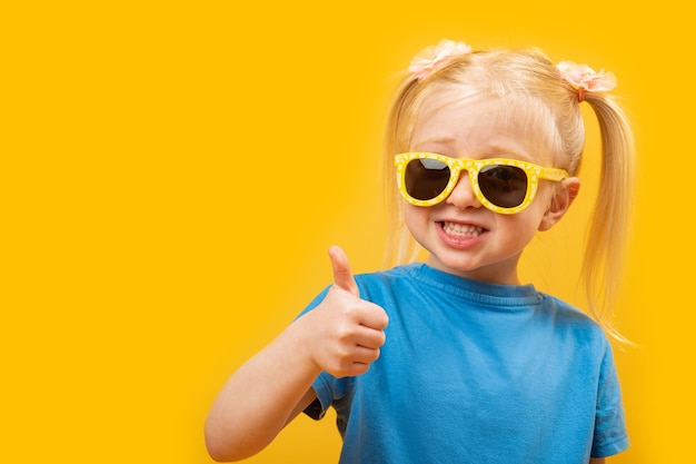 Cheerful preschool girl with blonde hair and two ponytails wears sunglasses Child shows thumbs up like gesture Copy space