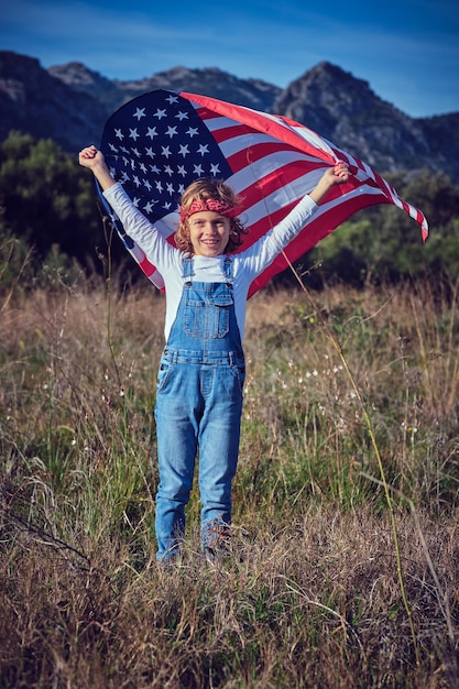 Cheerful preteen boy in casual clothes standing in meadow with waving American flag over head and looking at camera