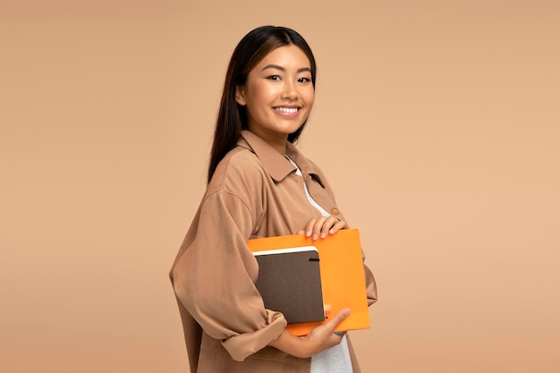 Cheerful pretty asian woman student holding books or paper organizers looking at camera