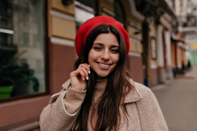 Cheerful pretty girl with closed great smile long dark hair in stylish red beret is smiling and posing against background of centre city