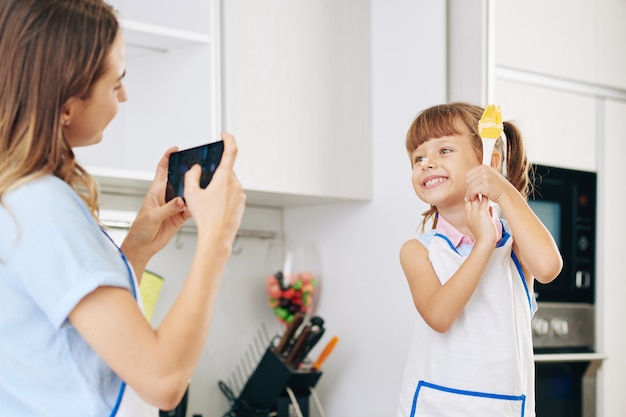 Cheerful pretty little girl with flour on her nose posing with silicone brush when mother taking photo on smartphone