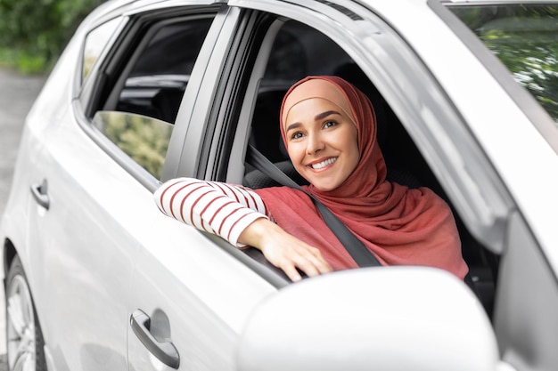 Cheerful pretty millennial middle eastern female in hijab ride in car with driver looks out open