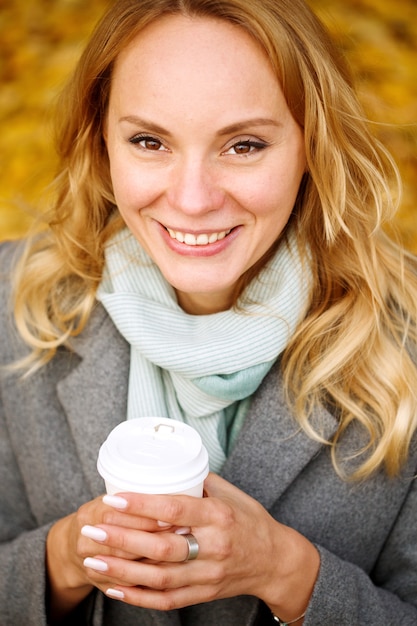 Cheerful pretty woman holding paper cup of coffee in autumn park looking at camera