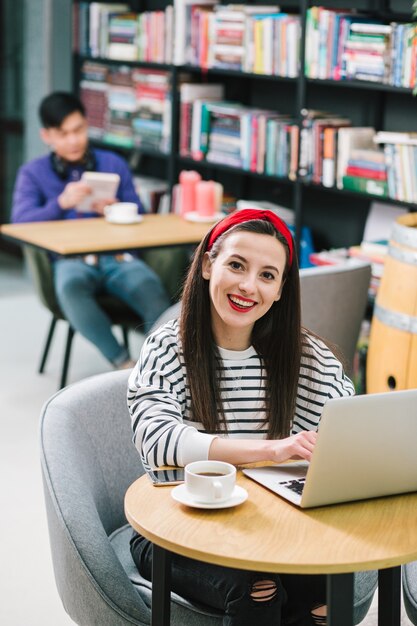 Cheerful pretty woman smiling while sitting with a laptop and a cup of coffee in a comfortable library