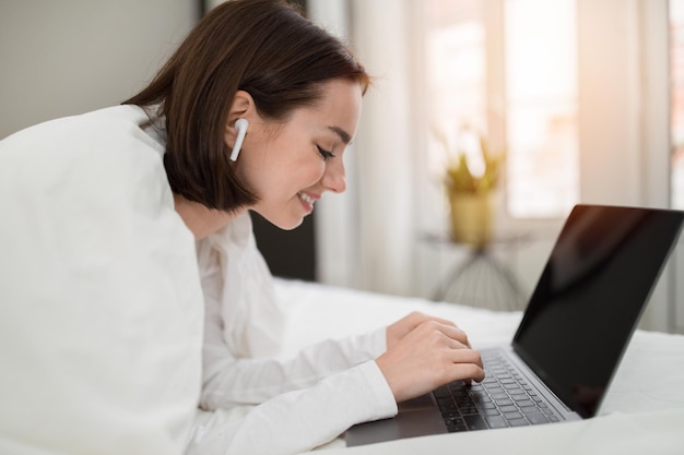 Cheerful pretty woman using laptop and earpods in bed mockup