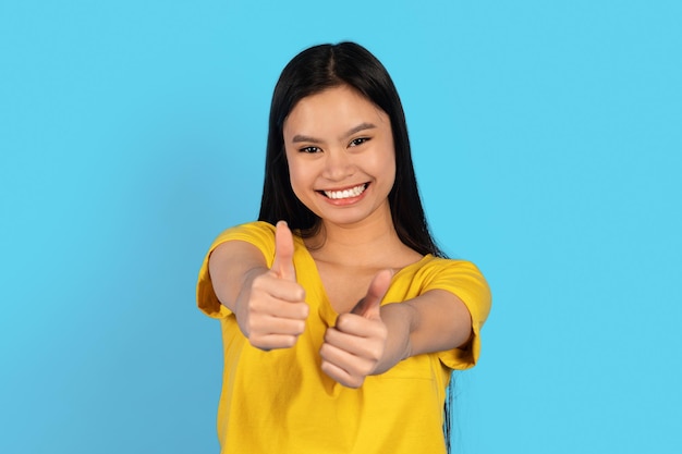 Cheerful pretty young chinese woman in yellow tshirt showing thumbs up gesture recommend ad