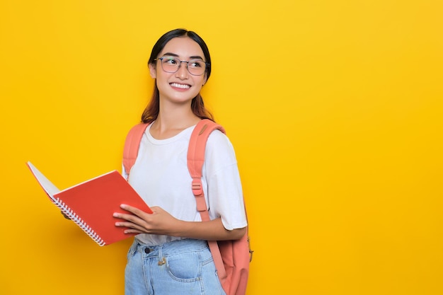 Cheerful pretty young student girl wearing backpack and eyeglasses holding books looking away at blank space isolated on yellow background