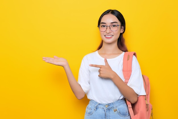 Cheerful pretty young student girl wearing backpack and eyeglasses presenting copy space on palm isolated on yellow background