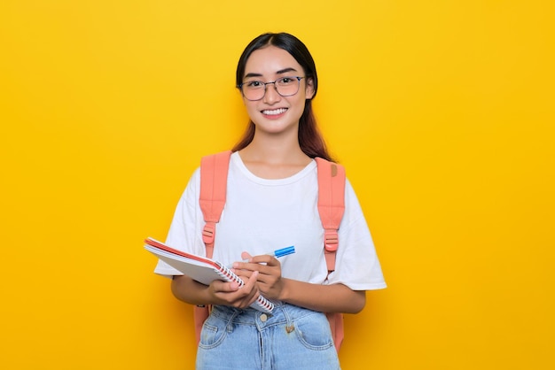 Cheerful pretty young student girl wearing backpack and eyeglasses writing in notebook isolated on yellow background