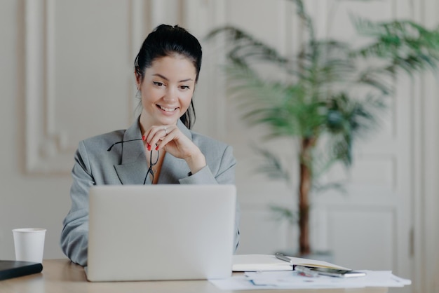 Cheerful professional woman searches laptop update removes glasses in formal office attire working remotely on blog ideas