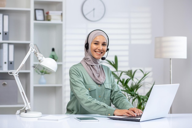 A cheerful professional woman wearing a hijab seated at a white desk using a laptop and headset in a