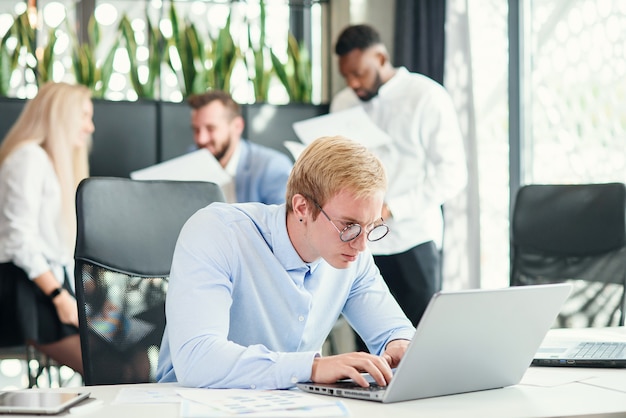 Photo cheerful purposeful male office worker with funny eyeglasses sits at his workplace and using laptop to solve financial tasks.