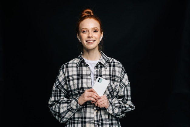 Cheerful red-haired young woman wearing wireless earphones holding phone and listening online music on isolated black background. Pretty redhead lady model emotionally showing facial expressions