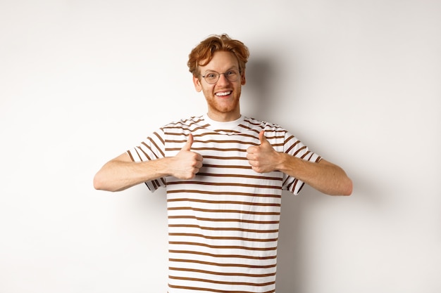 Cheerful redhead guy in nerdy glasses showing thumbs-up, smiling and saying yes, agree or like something, standing over white background.