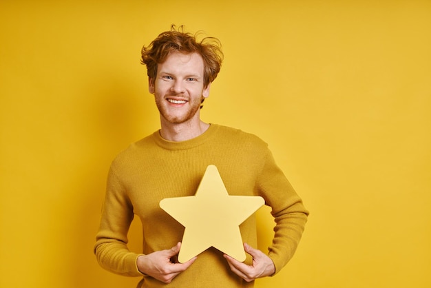 Cheerful redhead man holding star shape banner while standing against yellow background