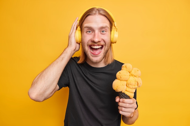 Cheerful redhead man smiles positively wears stereo headphones listens music has fun eats delicious ice cream dressed in black t shirt isolated over vivid yellow wall. Summer lifestyle