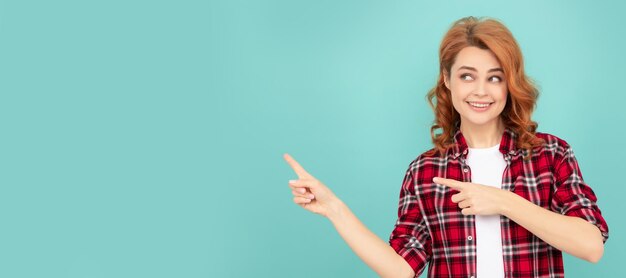 Cheerful redhead woman in checkered shirt point finger on blue background Woman isolated face portrait banner with mock up copyspace