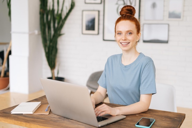 Cheerful redhead young woman is working on modern laptop computer at the desk at home office in cozy light room, looking at the camera. Concept of remote working on distance workplace.