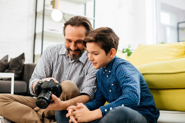 Cheerful relaxed Caucasian man sitting with his curious son and smiling while watching photos on the little screen of their modern camera