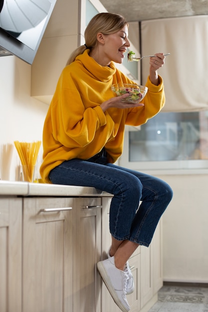 Cheerful relaxed woman on the kitchen countertop eating salad from the glass bowl and smiling