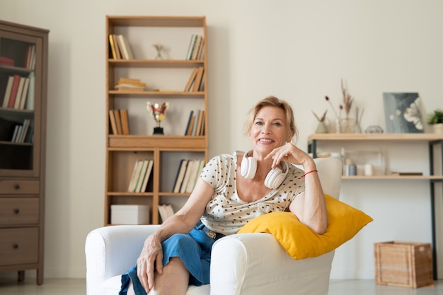 Cheerful restful middle aged female in homewear and headphones sitting in white leather comfortable armchair at home