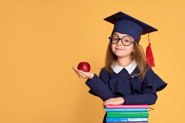 Cheerful schoolgirl in graduation outfit carrying apple while standing beside pile of textbooks