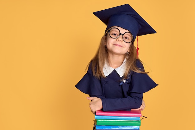 Cheerful schoolgirl in graduation outfit smiling while leaning on pile of textbooks