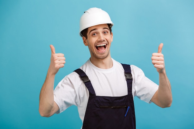 Cheerful screaming male builder in protective helmet showing thumbs up  over blue wall