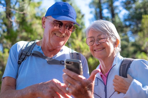 Cheerful senior couple of friends in mountain excursion looking mobile phone to check the right direction on the online map enjoying healthy lifestyle in nature