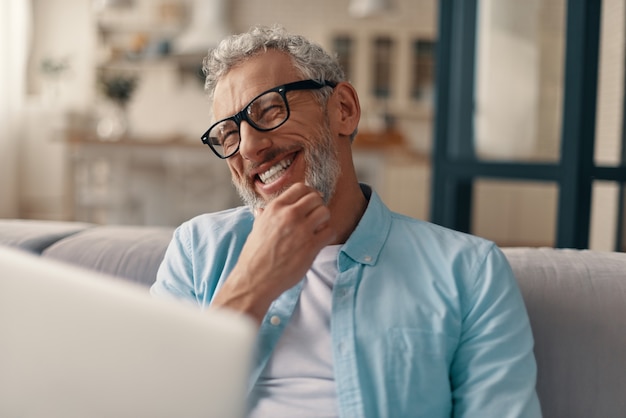 Cheerful senior man in casual clothing and eyeglasses using laptop and smiling while sitting on the sofa at home