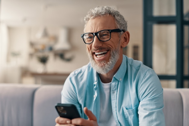 Cheerful senior man in casual clothing and eyeglasses using smart phone while sitting on the sofa at home