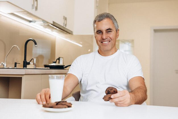 Cheerful senior man in white t-shirt has a glass of milk and chocolate cookies