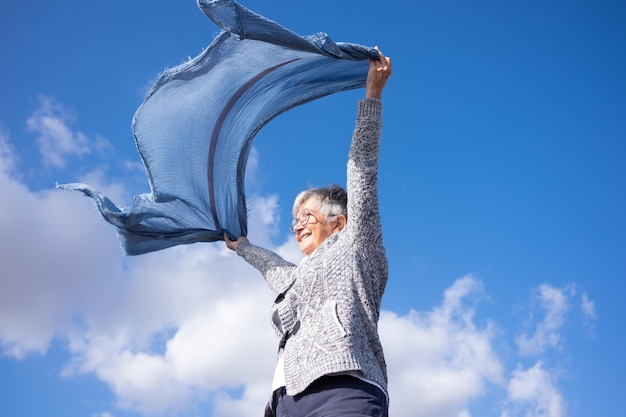 Cheerful senior woman standing outdoors with outstretched arms waving scarf in the blue sky enjoying freedom and vacation