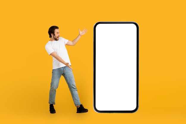 Cheerful shocked young european guy in white tshirt looks in surprise at huge phone with blank