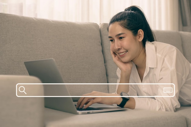 Cheerful smiling asian woman using laptop with search icon while sitting on couch in living room She uses her laptop for searching and enjoying shopping online