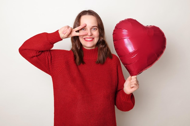 Cheerful smiling brown haired adult woman wearing red sweater holding heart shape balloon posing isolated over gray background enjoying festive event showing v sign near eyes rejoicing