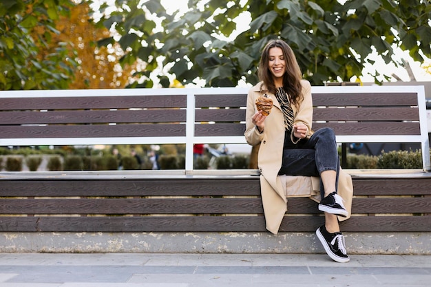 Cheerful smiling brunette girl sits on a bench with a croissant on the street The girl breaks off pieces eats a delicious croissant copy space